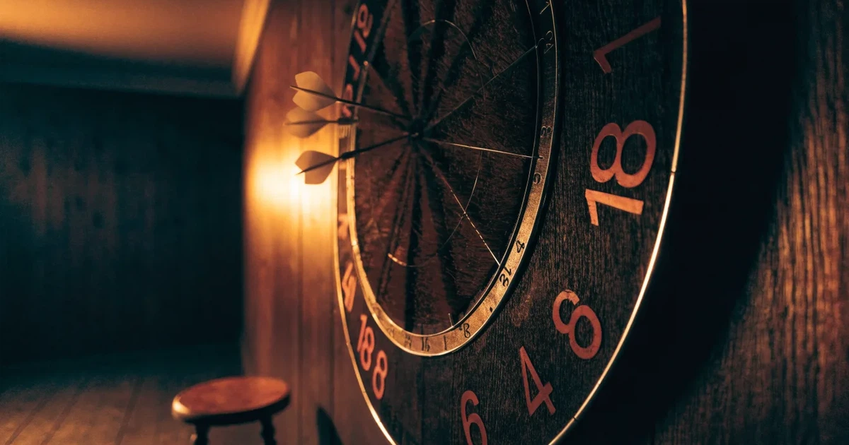 Eine Dartscheibe an einer Holzwand in einem schwach beleuchteten Raum, drei Pfeile stecken nahe der Mitte, warmes Licht von der Seite.